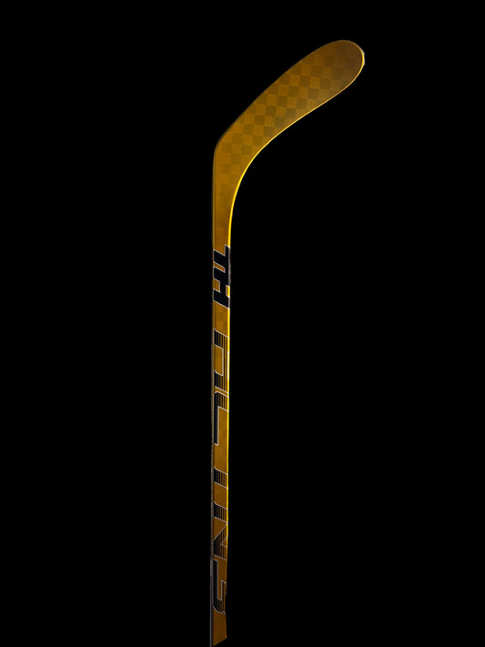 Hockey stick with gold and black design on a background of stacked hockey sticks. Taggs Hockey